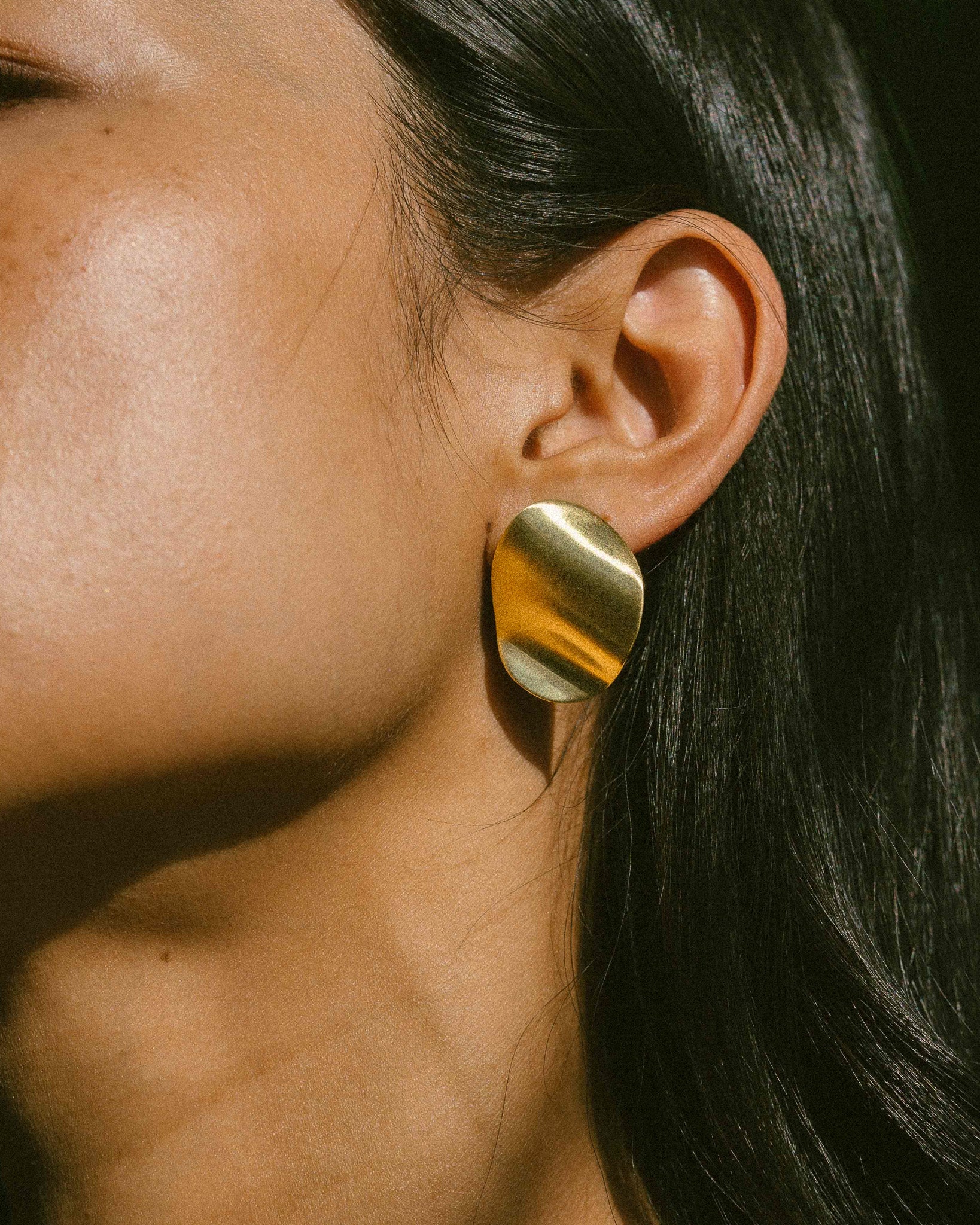 A model wearing a pair of clean line, pebble-shape inspired brass earrings.