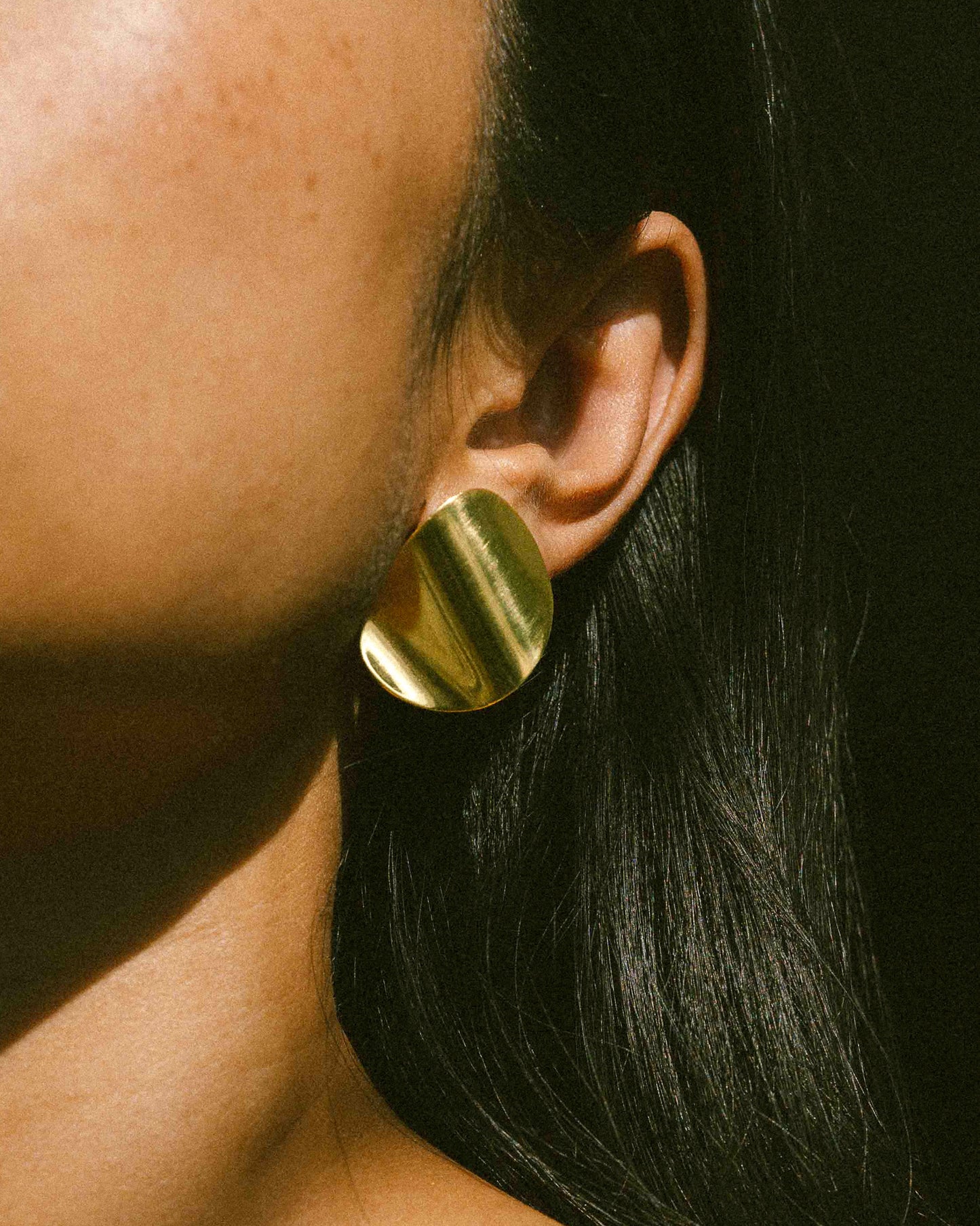 A model wearing a pair of clean line, pebble-shape inspired brass earrings.