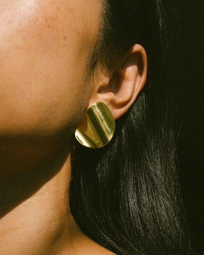 A model wearing a pair of clean line, pebble-shape inspired brass earrings.