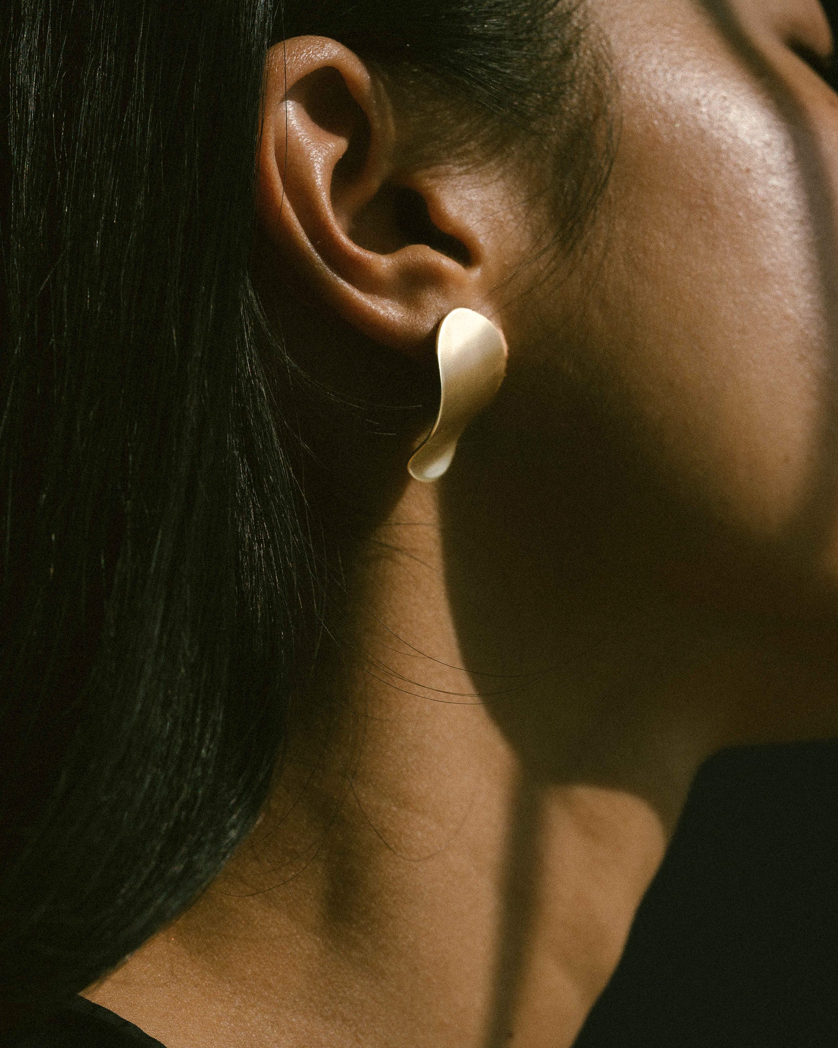 A model wearing a pair of clean line, pebble-shape inspired brass earrings.